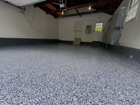 Empty garage interior with speckled gray epoxy floor, white walls, wooden ceiling beams, and a small high window