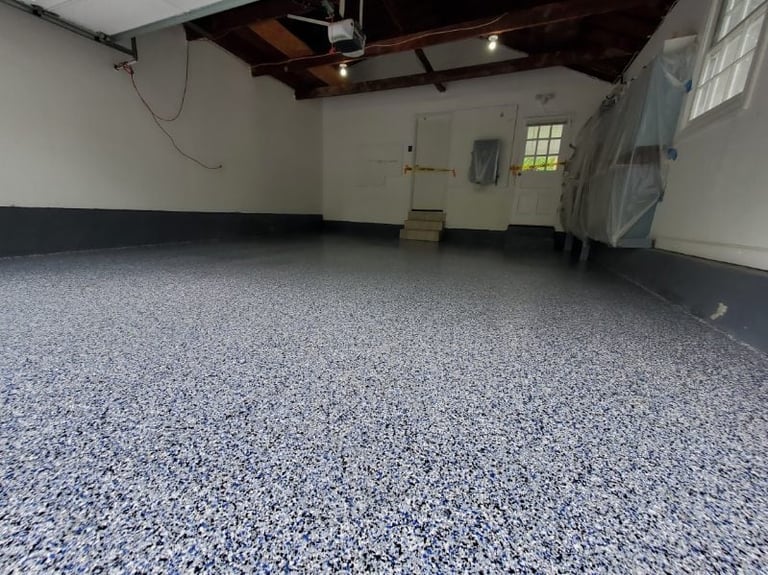 Empty garage interior with speckled gray epoxy floor, white walls, wooden ceiling beams, and a small high window