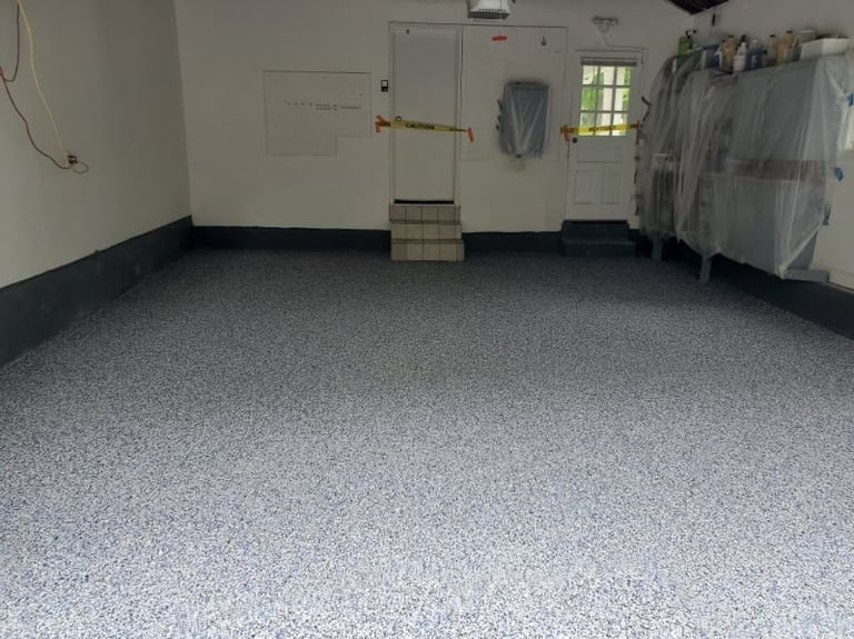 Empty garage interior with gray speckled epoxy floor, white walls, and storage items including hanging clothes and shelving.
