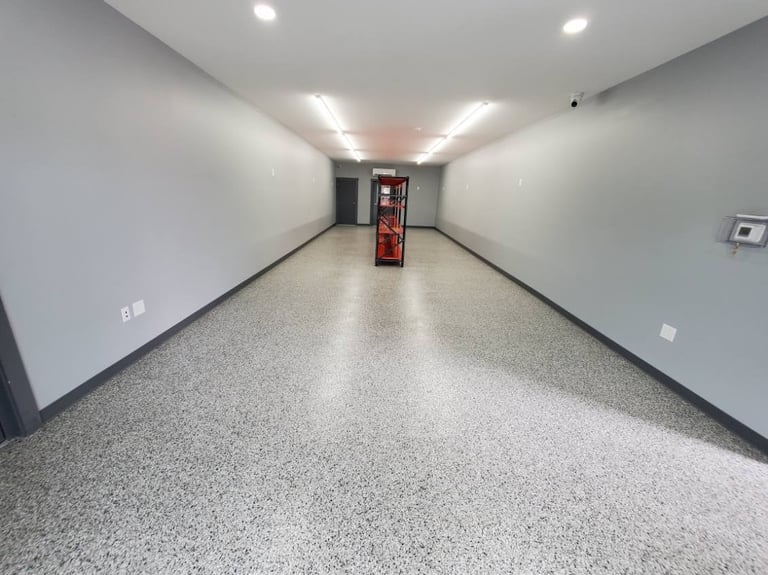 Empty hallway with white walls, speckled gray floor, ceiling lights, and a red fire extinguisher at the far end.